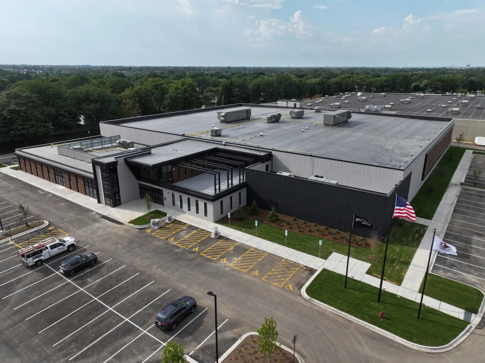 Aerial drone view of the arena's dark-panel and brick exterior, main glass entrance, and flat multi-sheet roof, with an American flag and a Spectate Group flag out front — Elk Grove Ice Arena