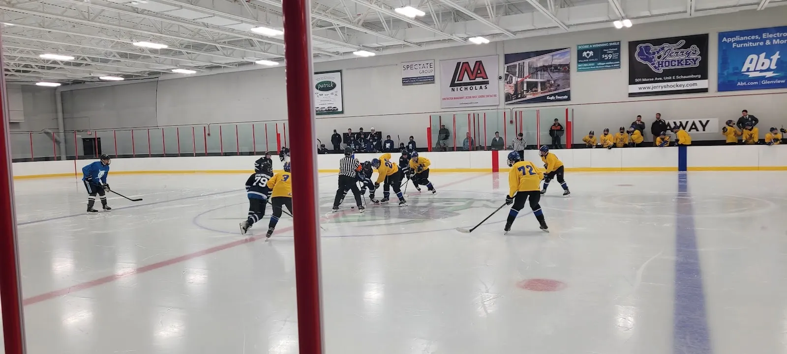 A faceoff near the far boards, players clustered across a wide sheet beyond a red steel post, sponsor boards and a long ceiling above. — Elk Grove Ice Arena