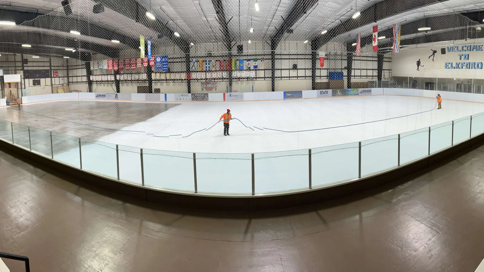 The Elkford Recreation Centre ice sheet being flooded and painted before the season, seen from the stands behind the glass, with flags and banners along the far wall and a Welcome to Elkford sign. — Elkford Recreation Centre