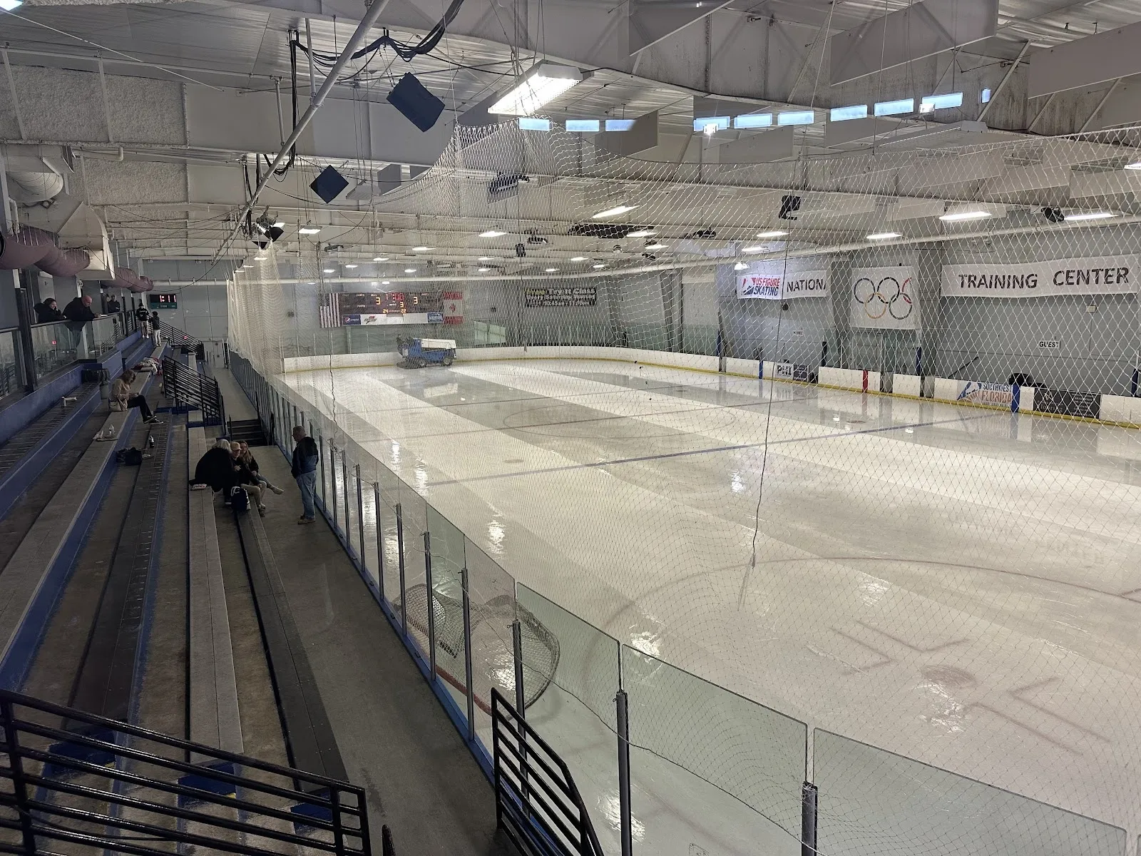 A long view down the sheet from the stands, empty white ice behind rink glass under a high pale roof with steel trusses. — Ellenton Ice and Sports Complex