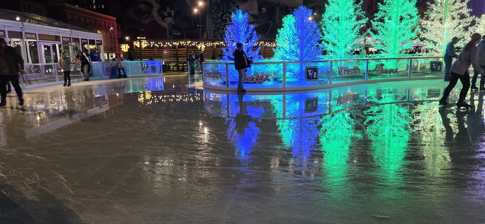 La pista de Elm Street Plaza de noche, con su hielo reflejando los árboles iluminados a lo largo de las vallas y patinadores a lo lejos en el borde — UC Health Ice Rink at Elm Street Plaza