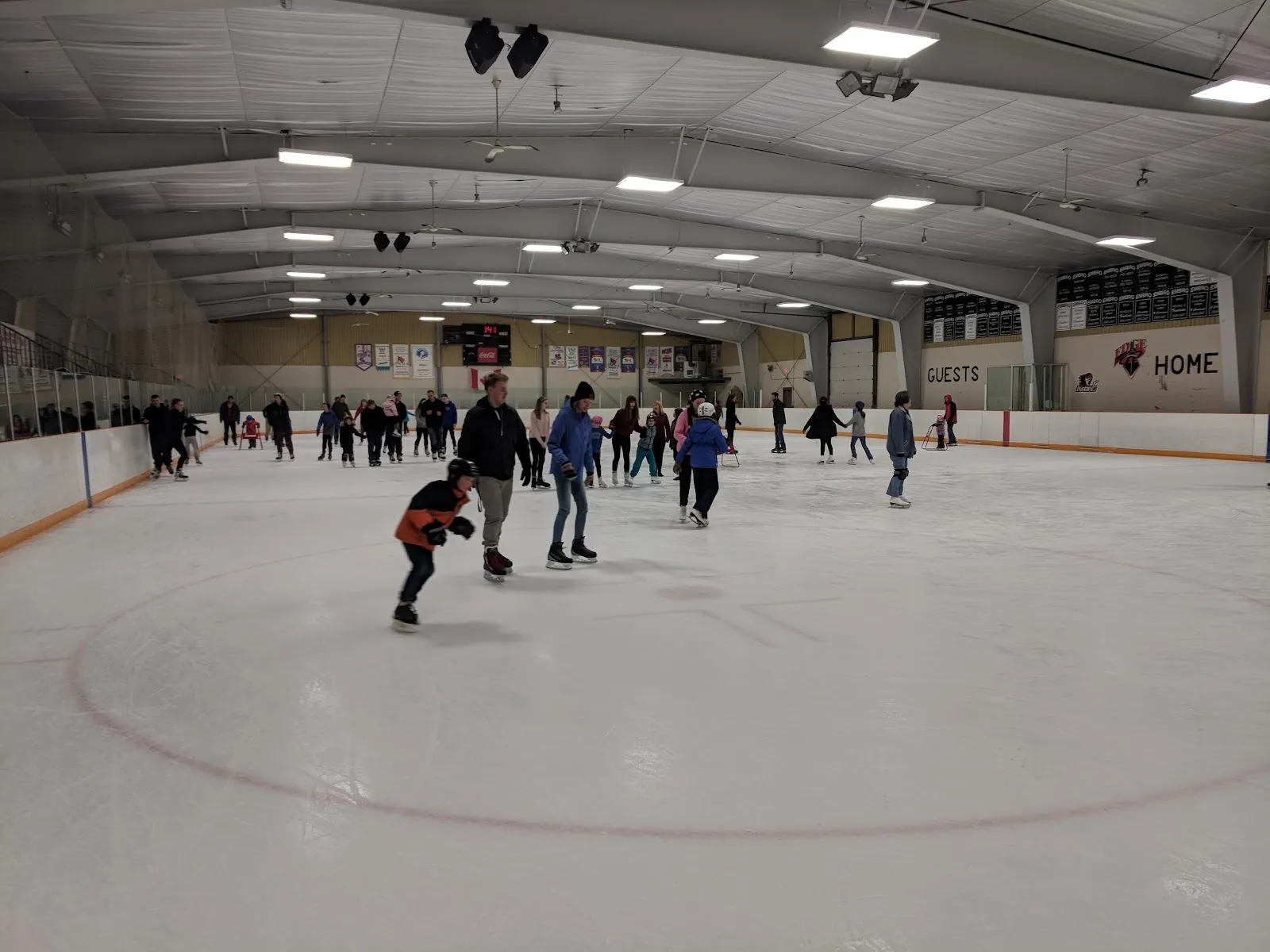 A public skating session on a wide sheet, skaters spread across the ice under a grey ceiling with steel girders and a long wall behind. — Embro-Zorra Community Centre