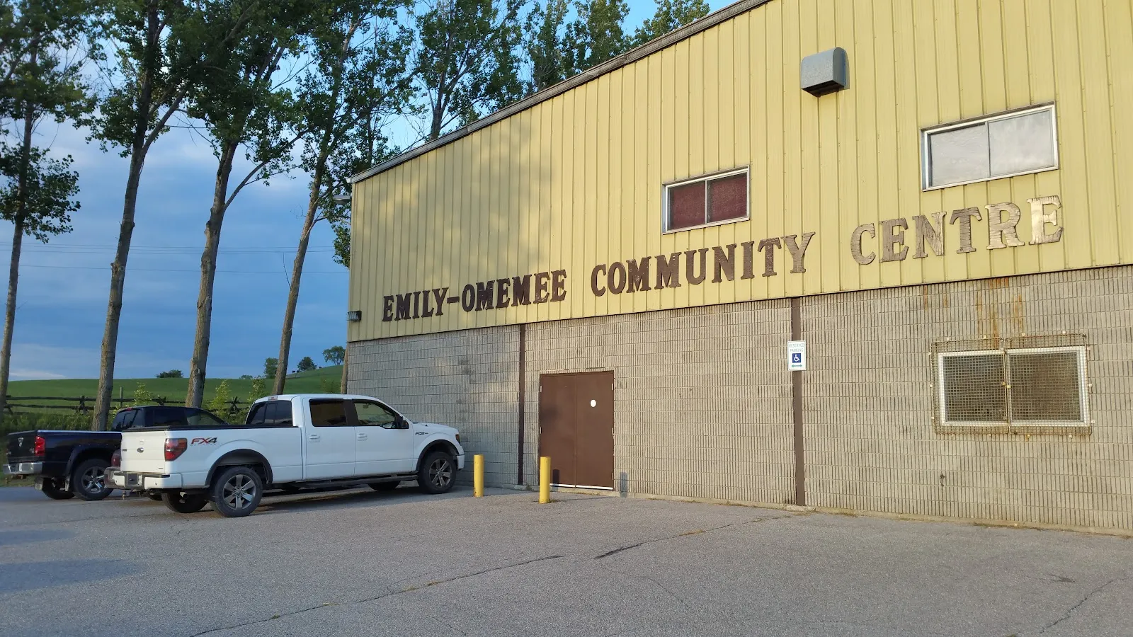 The tan metal arena wall with the Emily-Omemee Community Centre lettering and a side door off the lot. — Emily-Omemee Community Centre