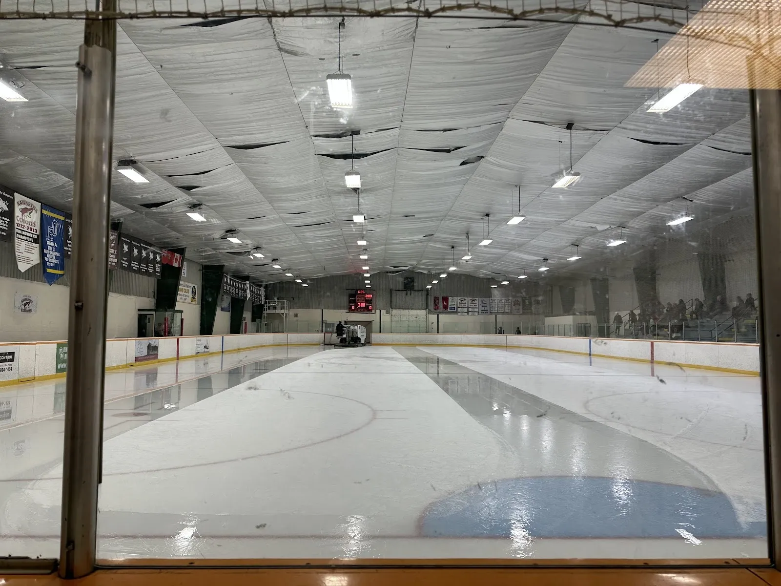 An empty sheet seen through netting from the end, the ice with a blue crease under a steel-trussed ceiling. — Emily-Omemee Community Centre