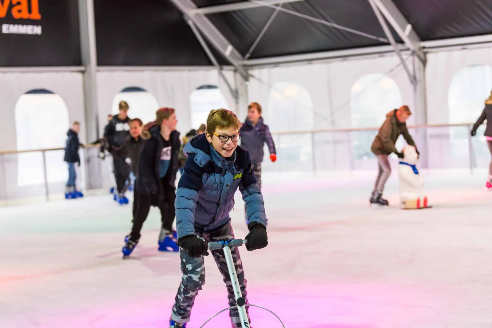 Kinderen schaatsen op de overdekte ijsbaan van Emmen on Ice, met het tentdak en de boarding erachter; schaatsers in actie, niemand poseert. – Emmen on Ice