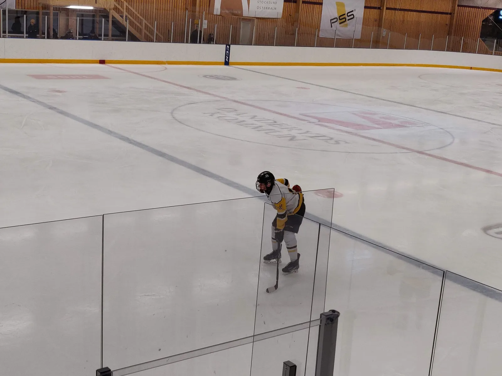 Isen i Enebybergs ishall med Danderyds kommuns logotyp i mitten av isen och en ensam spelare som väntar bakom plexiglaset vid sargen, med träväggar och sponsorbanderoller bakom – Enebybergs ishall