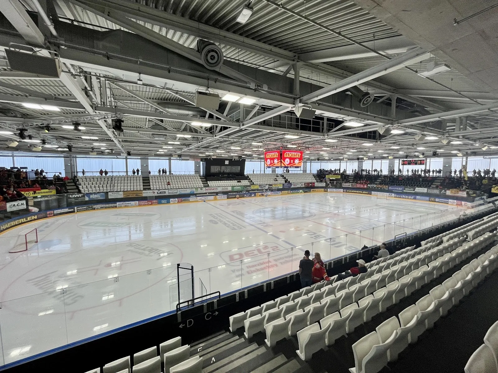 The ice sheet of the energie schwaben arena in Kaufbeuren from the stands, with the white seating, steel roof, scoreboard and board advertising; no one on the ice. — energie schwaben arena