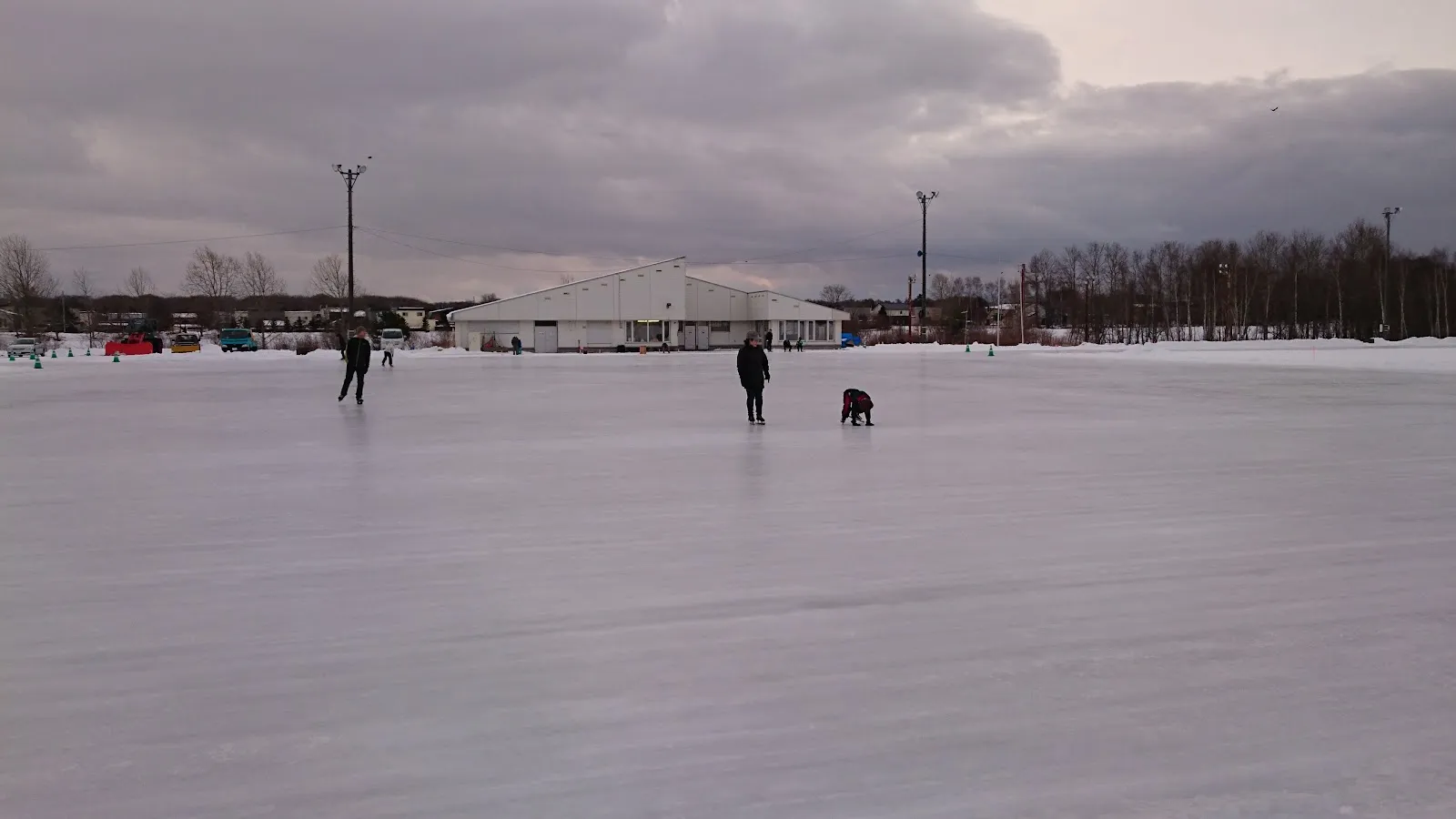 The wide ice sheet seen from the field edge, with a white warming hut and floodlight masts on the far side and three skaters in the distance. — Eniwa Citizen Skate Rink