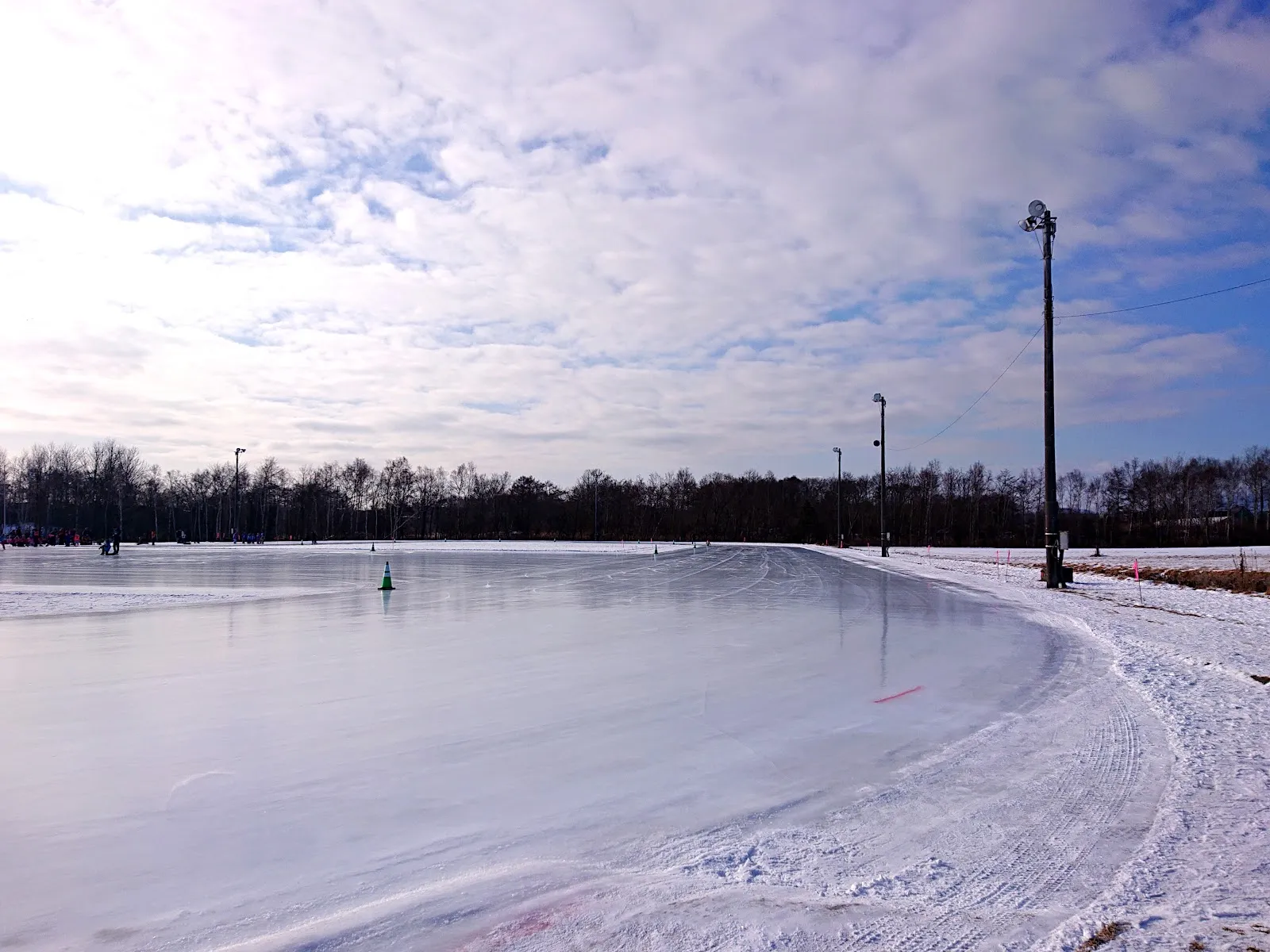 The curve of the outdoor skating oval's smooth ice on a winter afternoon, with floodlight masts along the edge, a line of bare trees behind and snow banked at the track side. — Eniwa Citizen Skate Rink