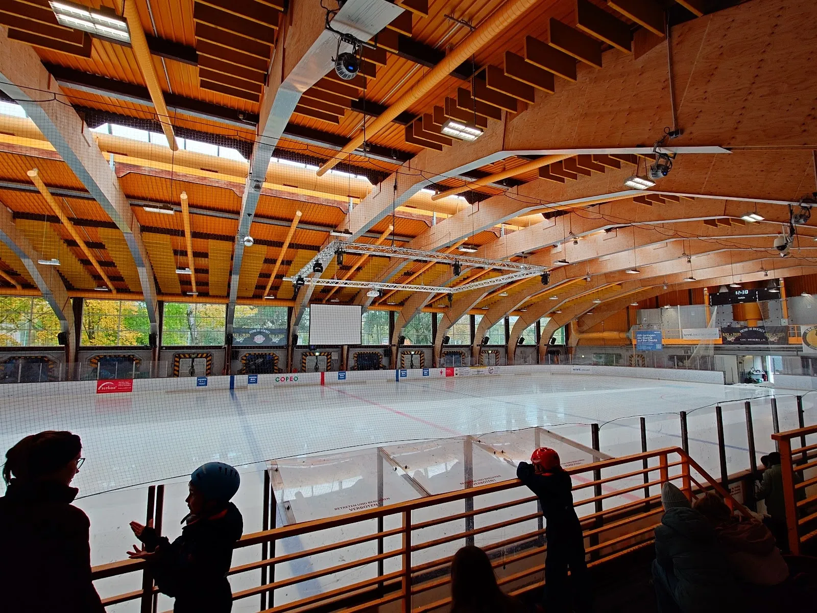 Die Eisfläche der enni.eiswelt in Moers vom Zuschauergeländer aus, mit den geschwungenen Holzbindern und großen Fenstern darüber und der Bande rund um das Eis. – enni.eiswelt