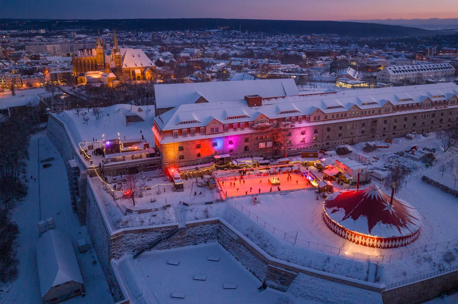 The Erfurter Winterberg rink on the Petersberg seen from above at dusk, the lit ice beside the fortress buildings with the city and cathedral behind it — Erfurter Winterberg (Eisbahn Petersberg)