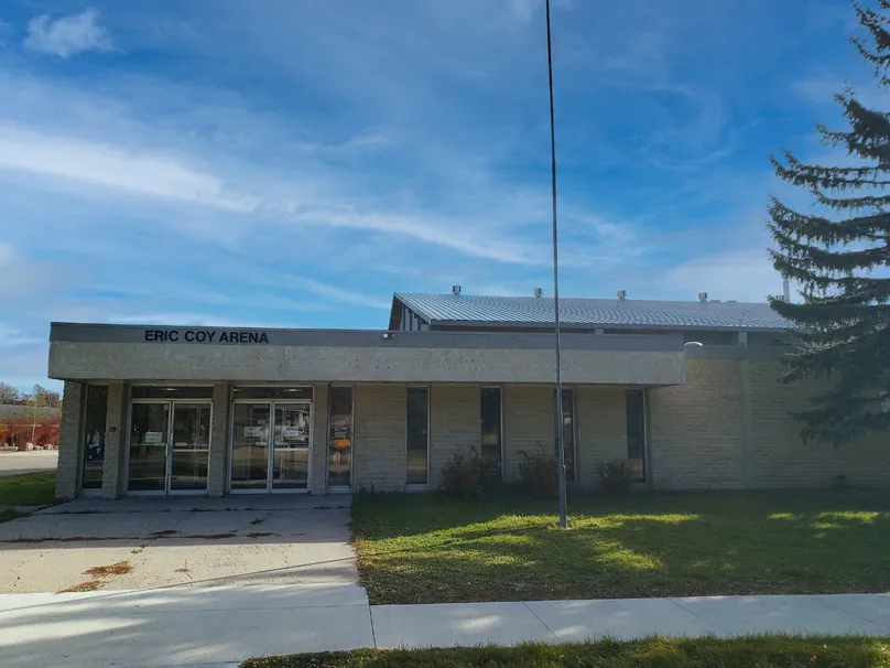 Beige brick building entrance with 'ERIC COY ARENA' lettering above glass double doors, angled metal roof visible behind, flagpole and sidewalk in front. — Eric Coy Arena