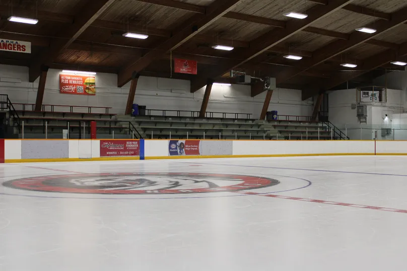 Full ice sheet with a team logo painted at centre ice, empty wooden-raftered stands and rink boards visible, no people on the ice. — Eric Coy Arena
