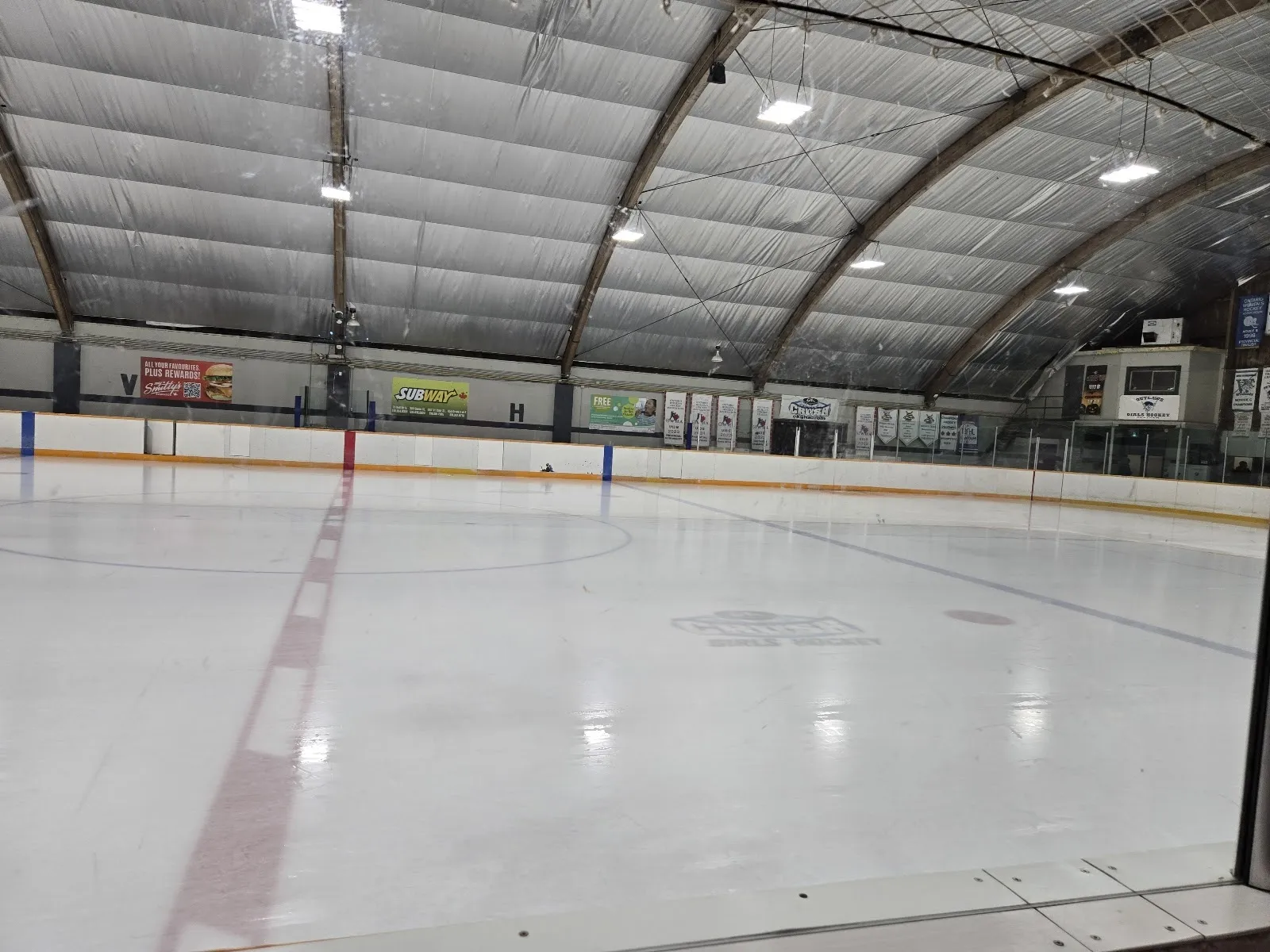 Una pista amplia y vacía a lo largo de las vallas, con hielo blanco y una línea roja bajo un techo arqueado de acero verde. — William K. Erickson Arena