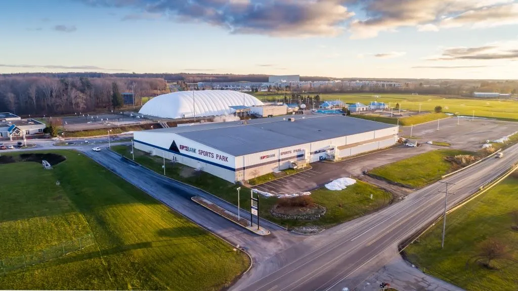 Aerial view of the low white metal building with "ERIEBANK SPORTS PARK" painted on the side wall, parking lot and access road in front, the white dome fieldhouse visible behind it at dusk — Erie Sports Park (LECOM Sports Park)