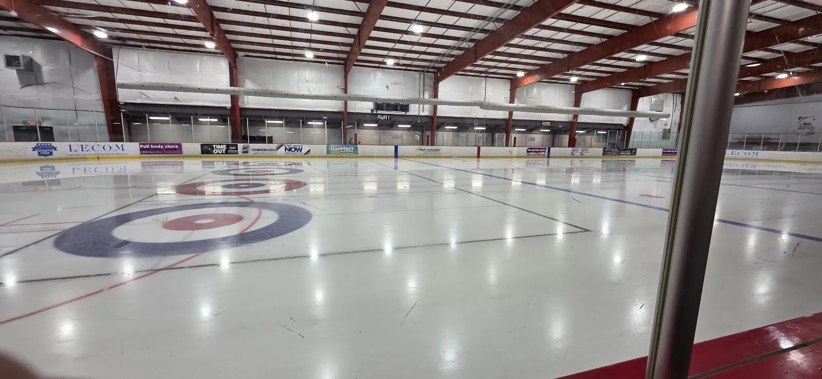 An empty sheet with a blue and red circle on the ice, red boards and a pale steel-trussed roof seen through the glass. — Erie Sports Park (LECOM Sports Park)