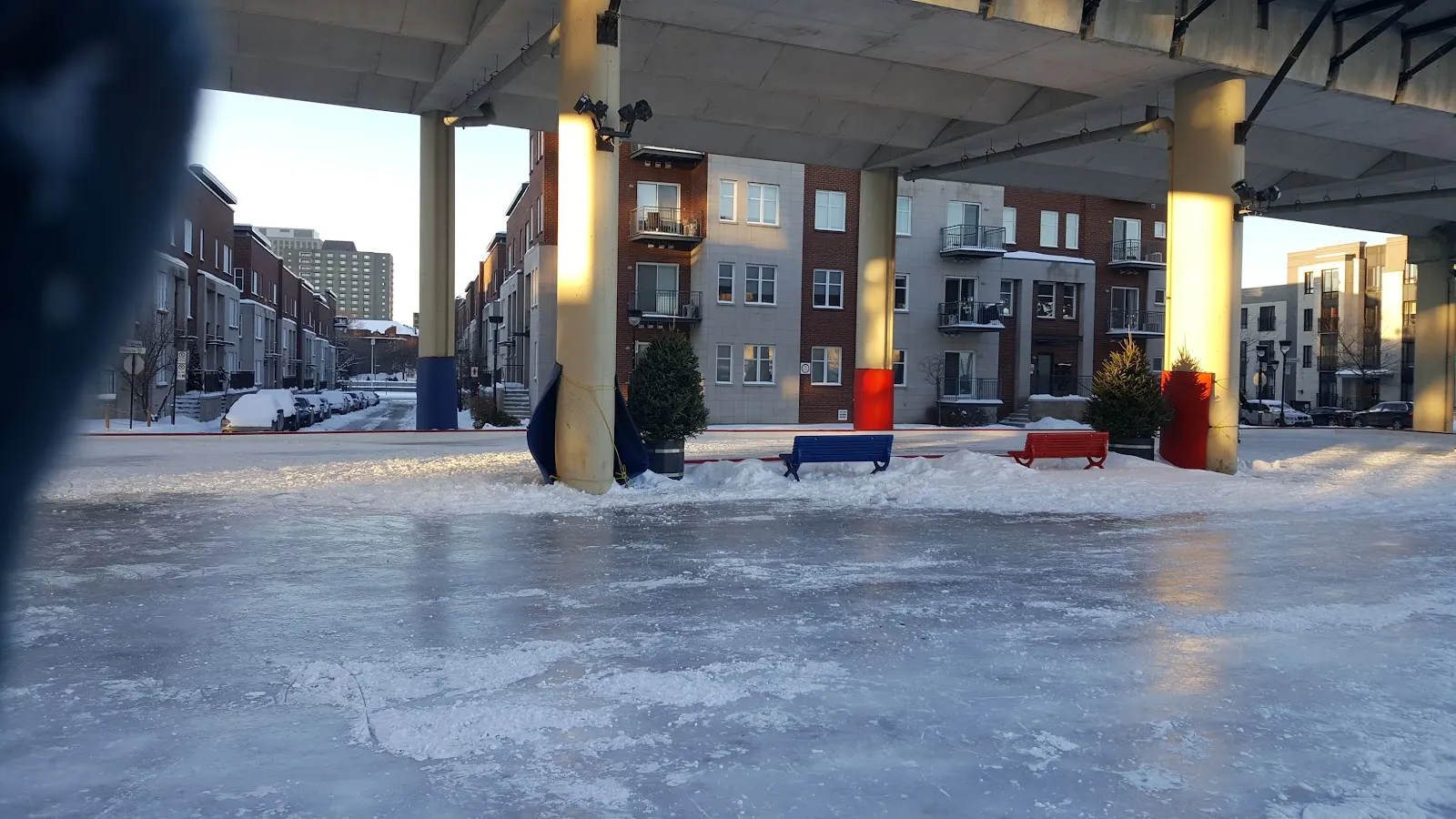 The ice of the Glacis rink at Espace Faubourg Québec in winter, under the covered structure with benches and brick apartment buildings behind — Espace Faubourg Québec Outdoor Rink