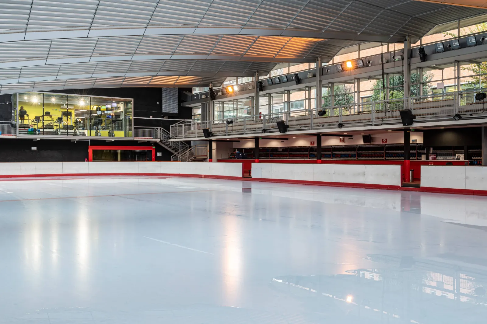 The empty ice sheet with the red-and-white boards, the spectator mezzanine and the glass-fronted fitness room behind, under a curved roof with glazing. — Patinoire Pailleron
