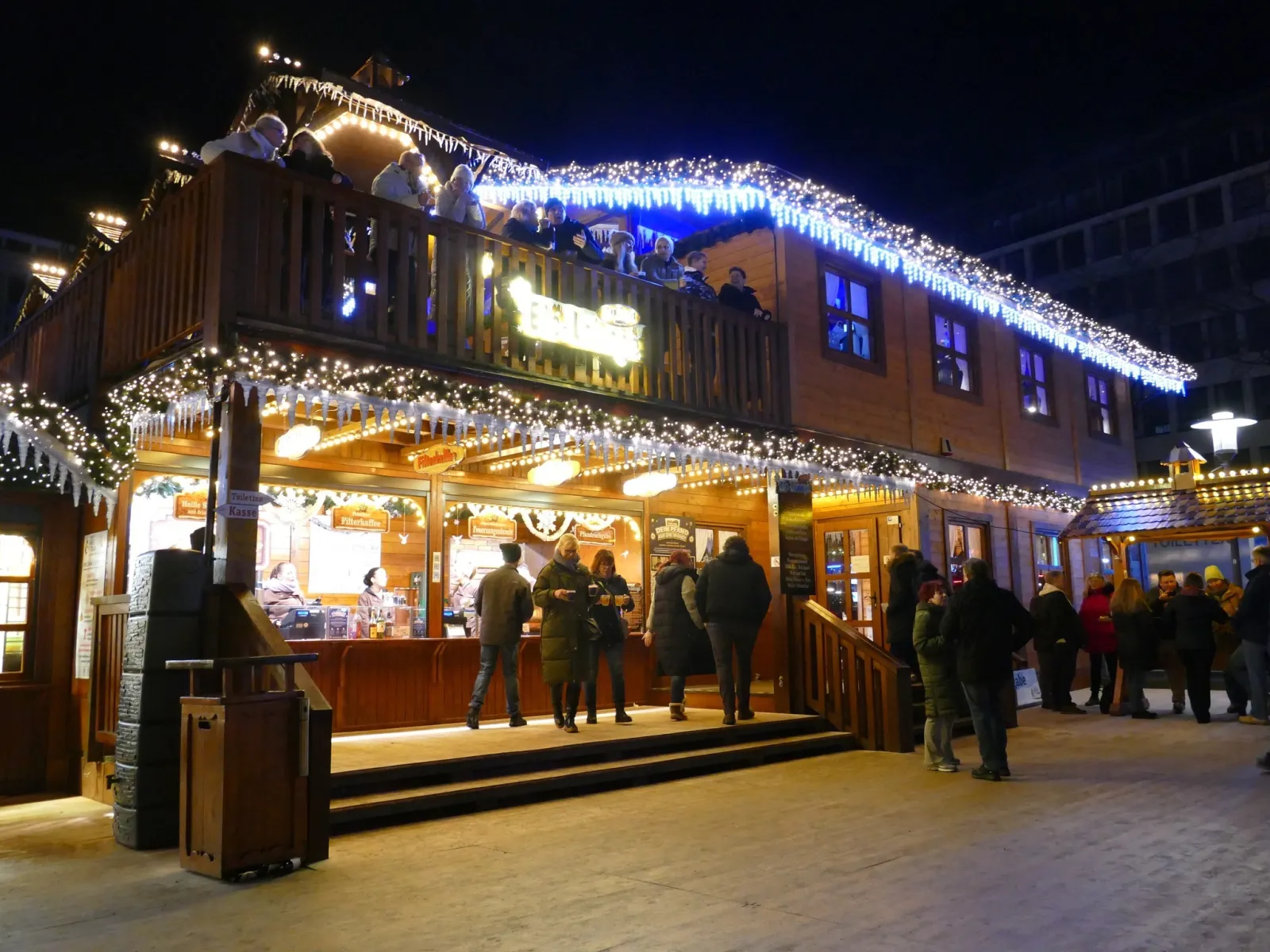 The two-storey Eis-Lounge of the Essener Wintertraum on the Kennedyplatz at night, wrapped in fairy lights, with guests at the ground-floor counter. — Essener Wintertraum