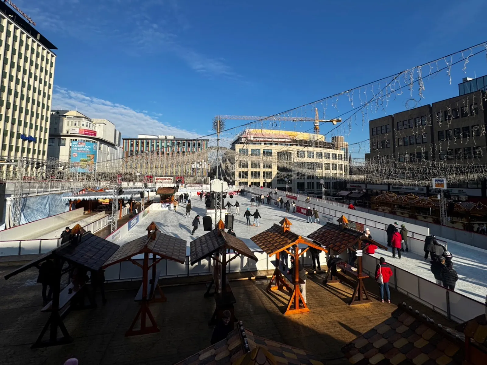 The Essener Wintertraum rink on the Kennedyplatz seen from the upper level on a sunny day, with skaters at a distance on the long ice and the city buildings behind. — Essener Wintertraum