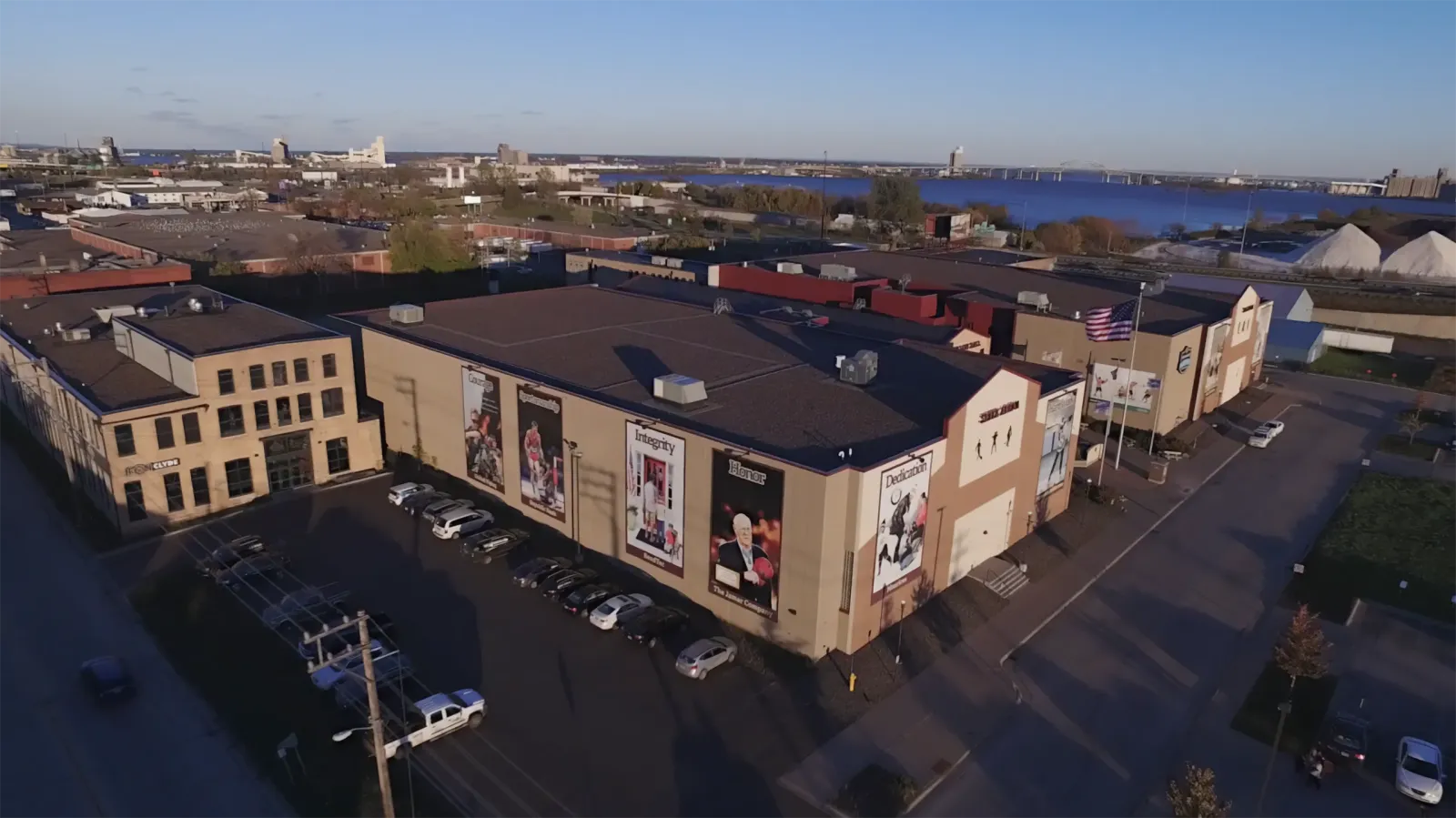 Aerial view of the Heritage Center building (Sill Arena side) showing its Clyde Iron Works-themed facade with painted murals, the American flag, and the parking lot, with the St. Louis River and downtown Duluth in the background. — Essentia Duluth Heritage Center