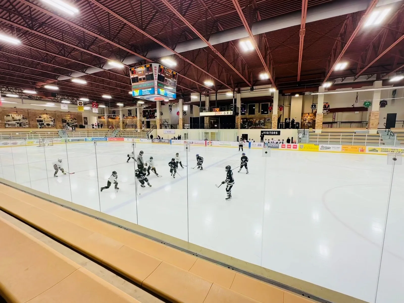 A game seen along the boards, players spread across the ice under a roof of dark red timber beams with a pale far wall. — Essentia Duluth Heritage Center