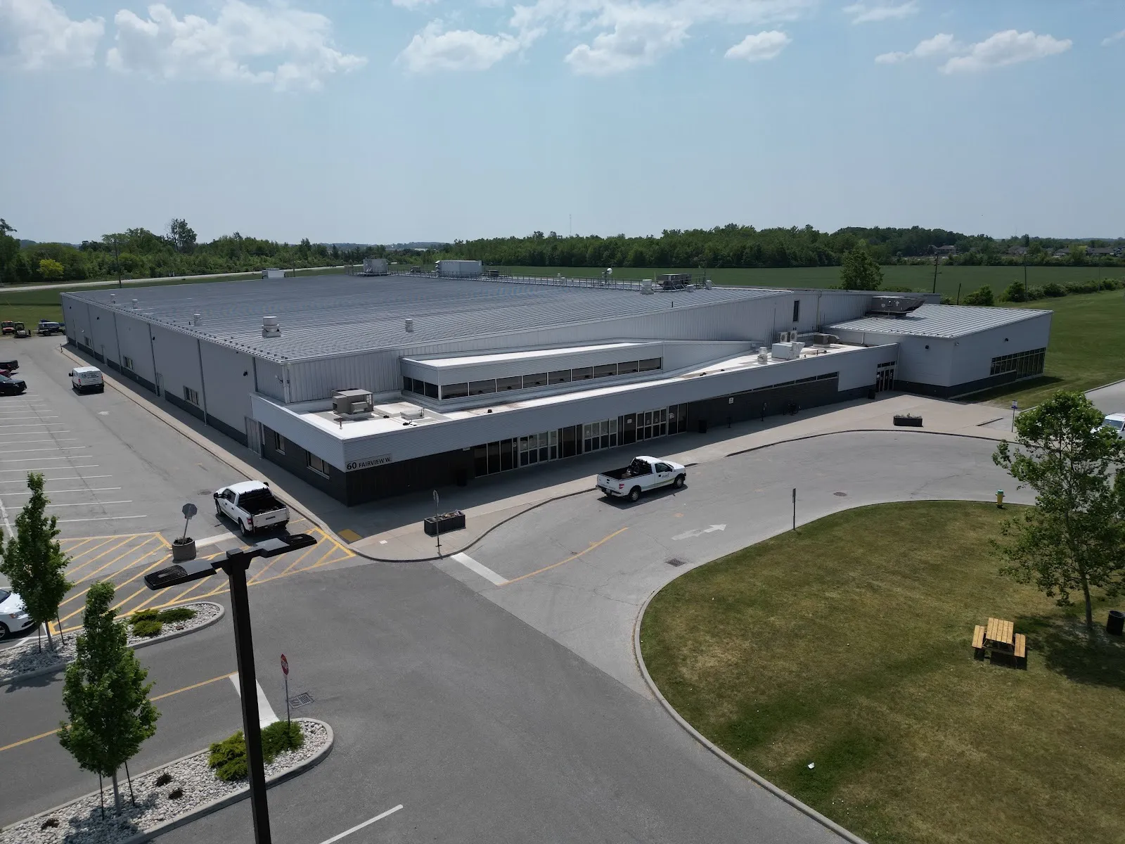 El Essex Centre Sports Complex desde arriba: un edificio gris, largo y bajo, con un bloque de entrada de vidrio, y estacionamiento y un camino de acceso curvo al frente. — Essex Centre Sports Complex