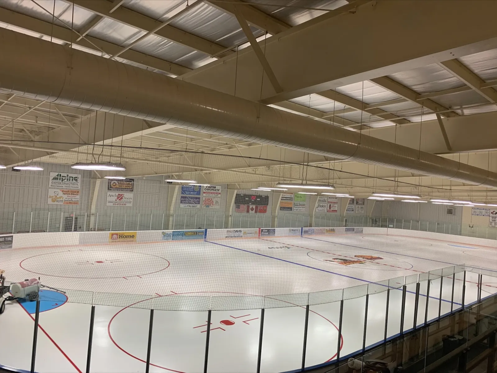 Una pista vacía vista desde encima del cristal: hielo blanco recién hecho con líneas rojas y azules, anuncios de patrocinadores a lo largo del lado lejano y un techo de acero color beige con lámparas colgantes. — Essex Centre Sports Complex