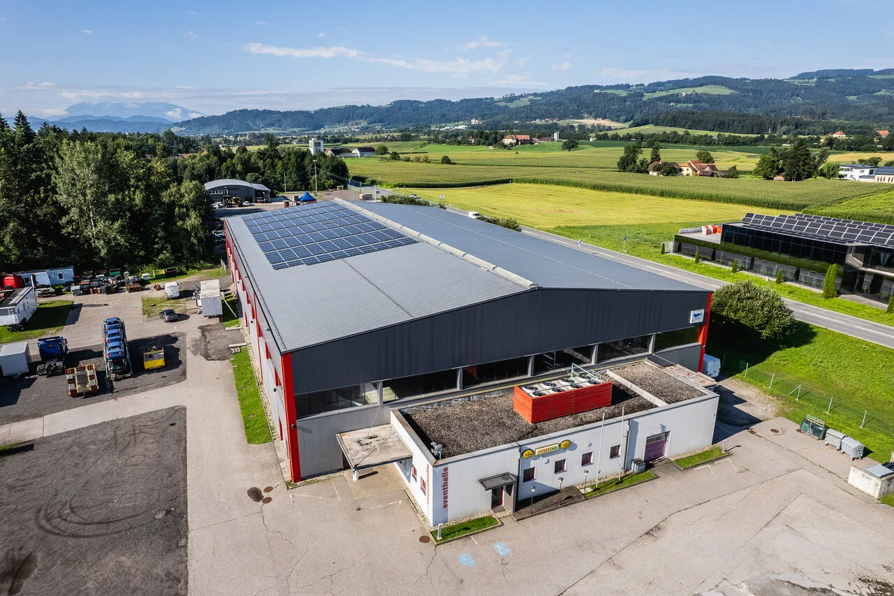 Die Eventhalle Wolfsberg von oben: eine Halle mit Solardach, davor ein Parkplatz und Wiesen, dahinter die Berge – Eishalle Wolfsberg (Eventhalle Kleinedling)