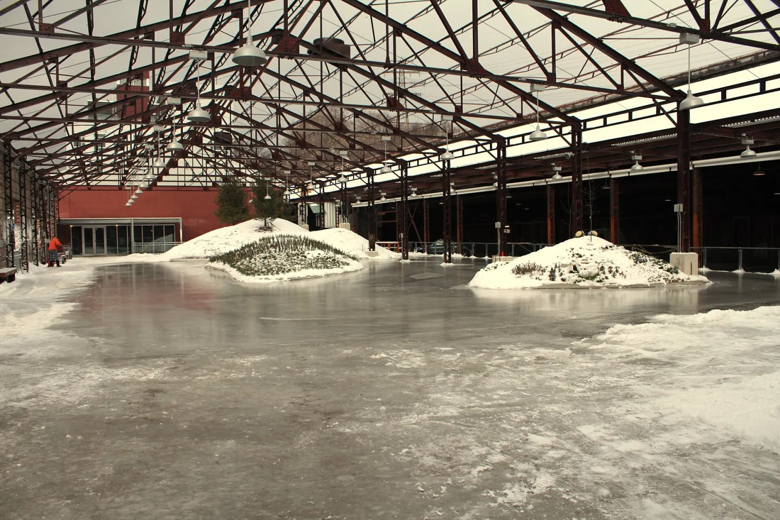 The Evergreen Brick Works skating rink inside the old brick-works shed, a sheet of ice with two snow piles under the red steel roof trusses. — Evergreen Brick Works Skating Rink
