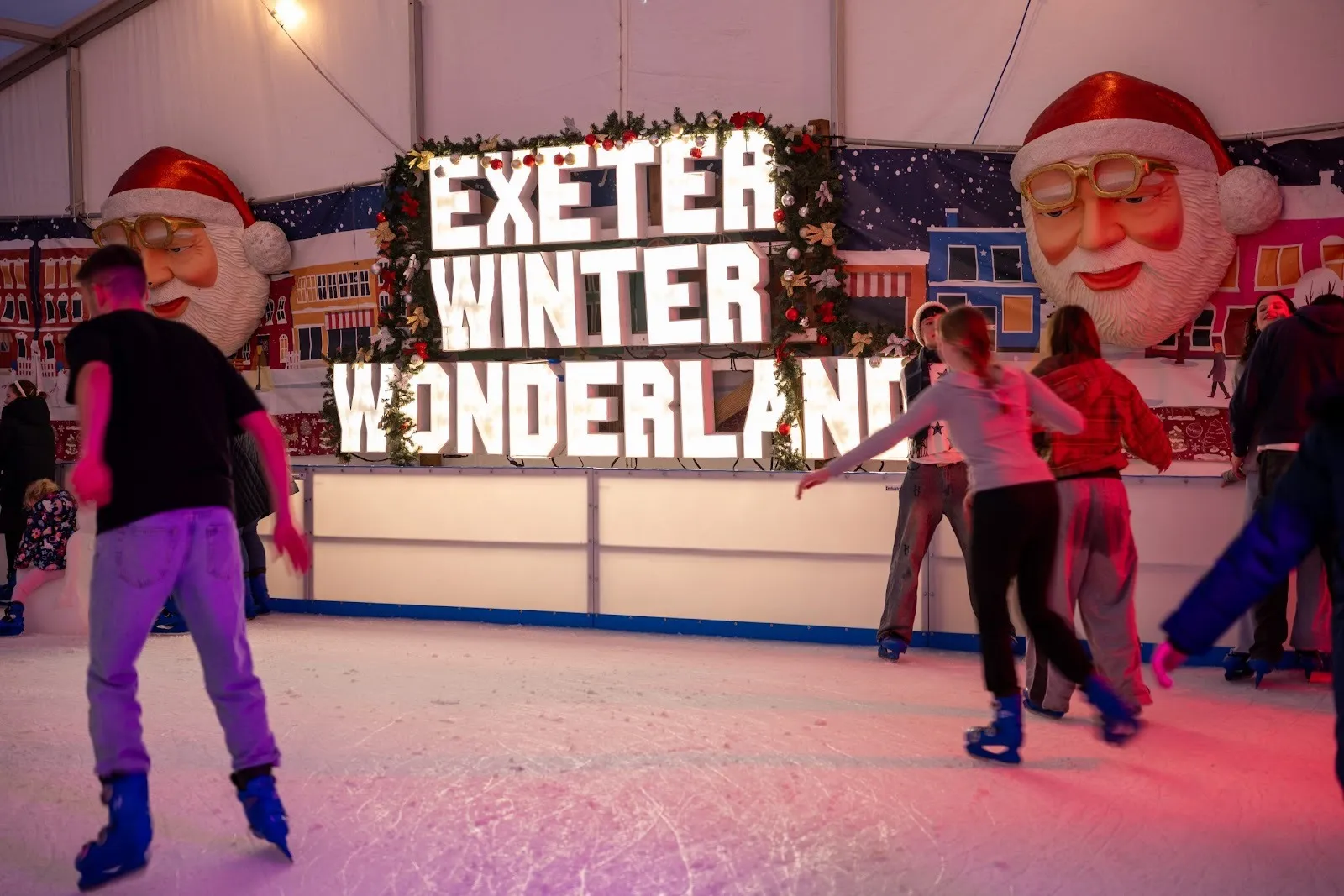 Skaters on the Exeter Winter Wonderland rink in front of a lit sign flanked by two giant Santa figures. — Exeter's Winter Wonderland Ice Rink