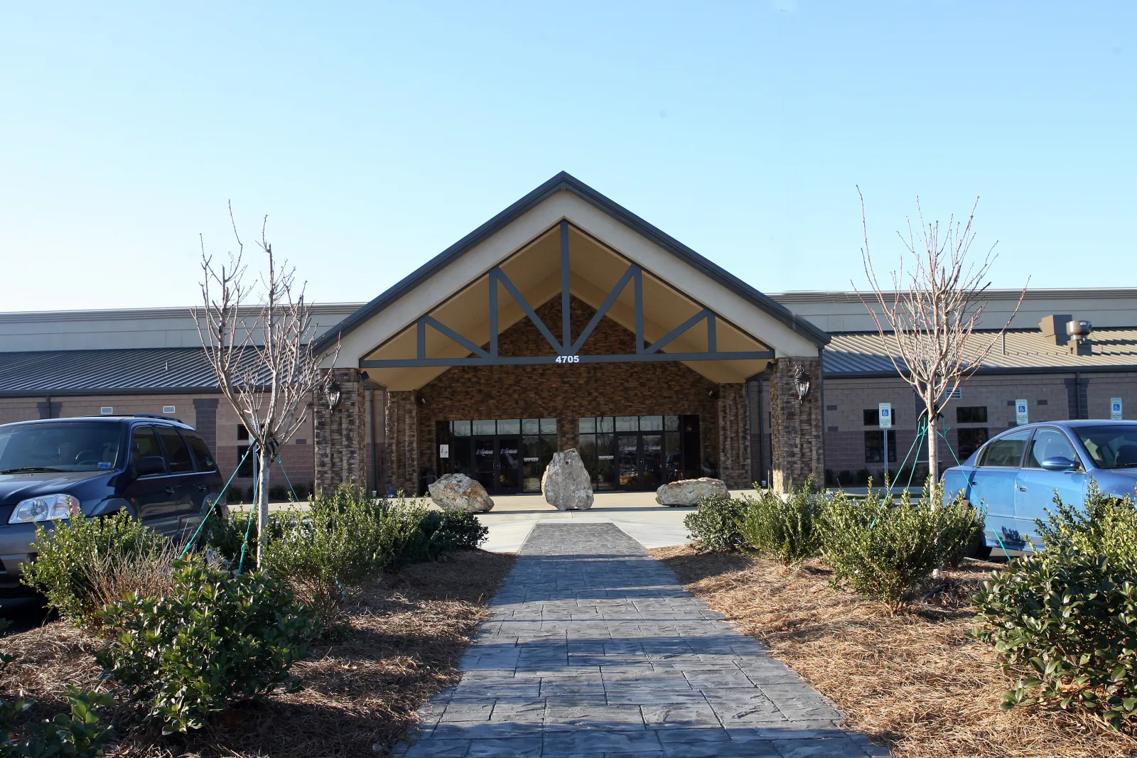 Extreme Ice Center's stone-and-timber front entrance under a gabled canopy marked '4705', flanked by young trees and a parking lot with accessible spaces, on a clear day. — Extreme Ice Center