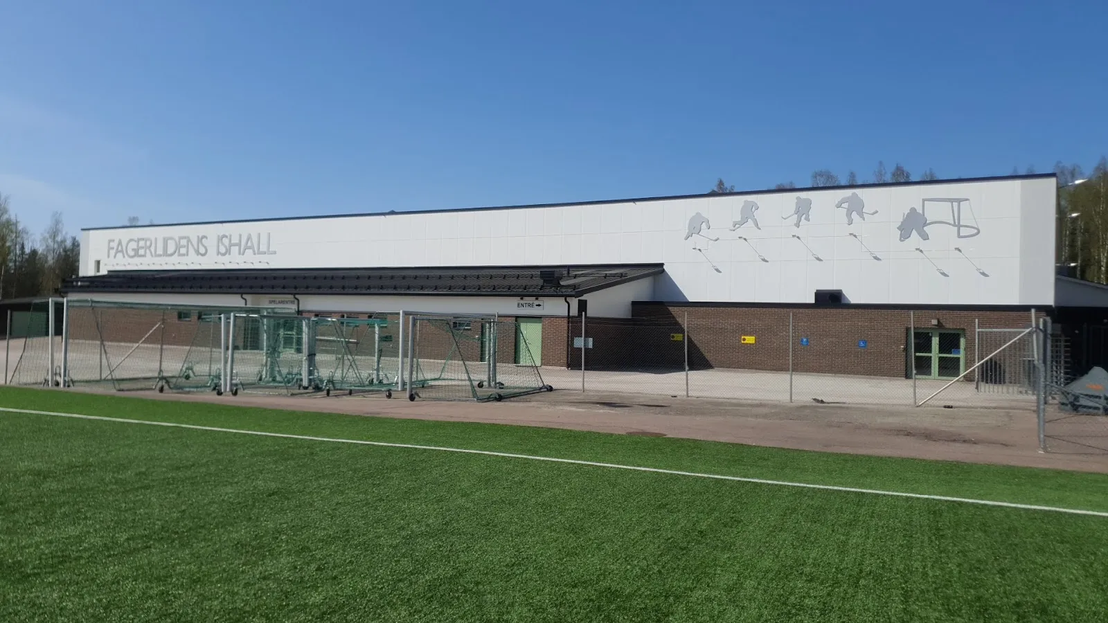 Den låga vita byggnaden Fagerlidens ishall med sin namnskylt, sedd över fotbollsplanen – Fagerlidens ishall