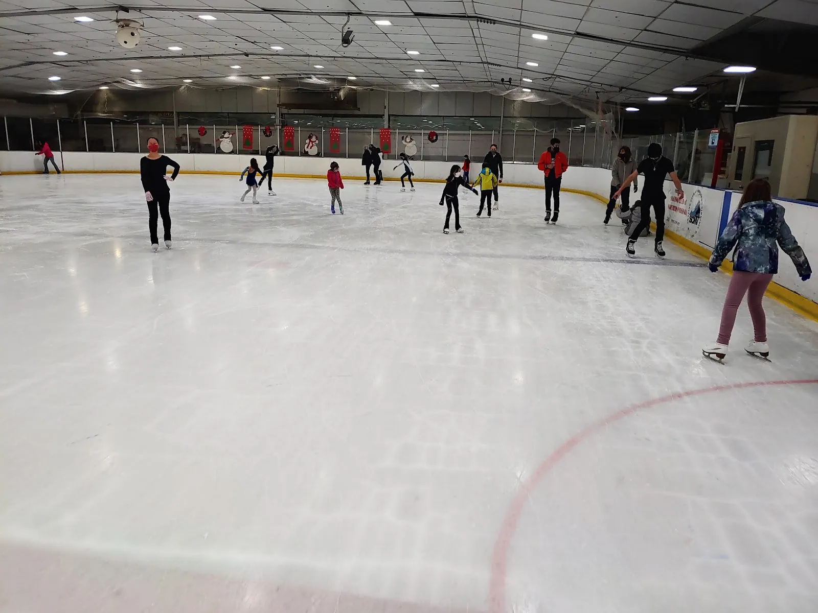 Una sesión pública en el hielo con patinadores repartidos a lo largo de las vallas del fondo bajo un alto techo de acero, con una línea roja curvándose en primer plano. — Fairfax Ice Arena