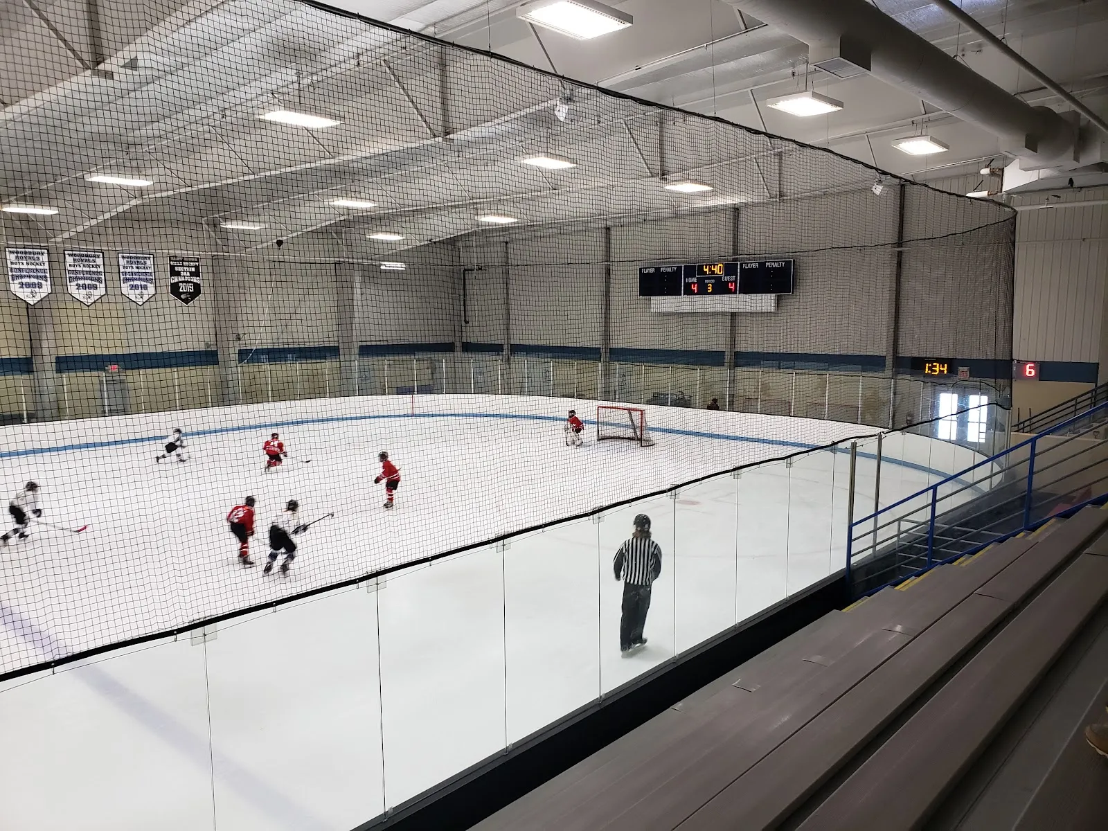 A hockey practice on the indoor ice sheet seen from the tiered seating, with players spread around the near end, a net at the far end and glass above the boards under a steel-truss roof. — M Health Fairview Sports Center