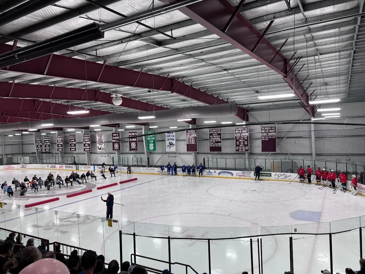 A game seen from the stands, players spread across the ice with a red-trussed ceiling above and green and white banners along the far wall. — Falmouth Ice Arena