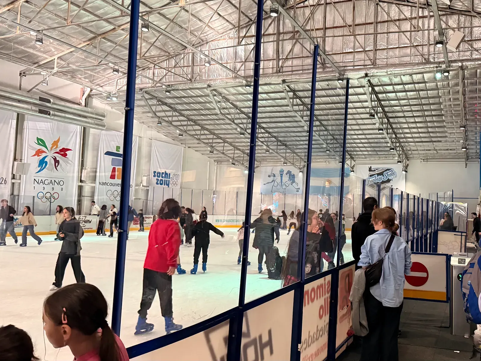 La amplia pista bajo el techo de acero durante una sesión pública, con banderas olímpicas a lo largo del muro del fondo y patinadores repartidos por el hielo — Fantasy Skate