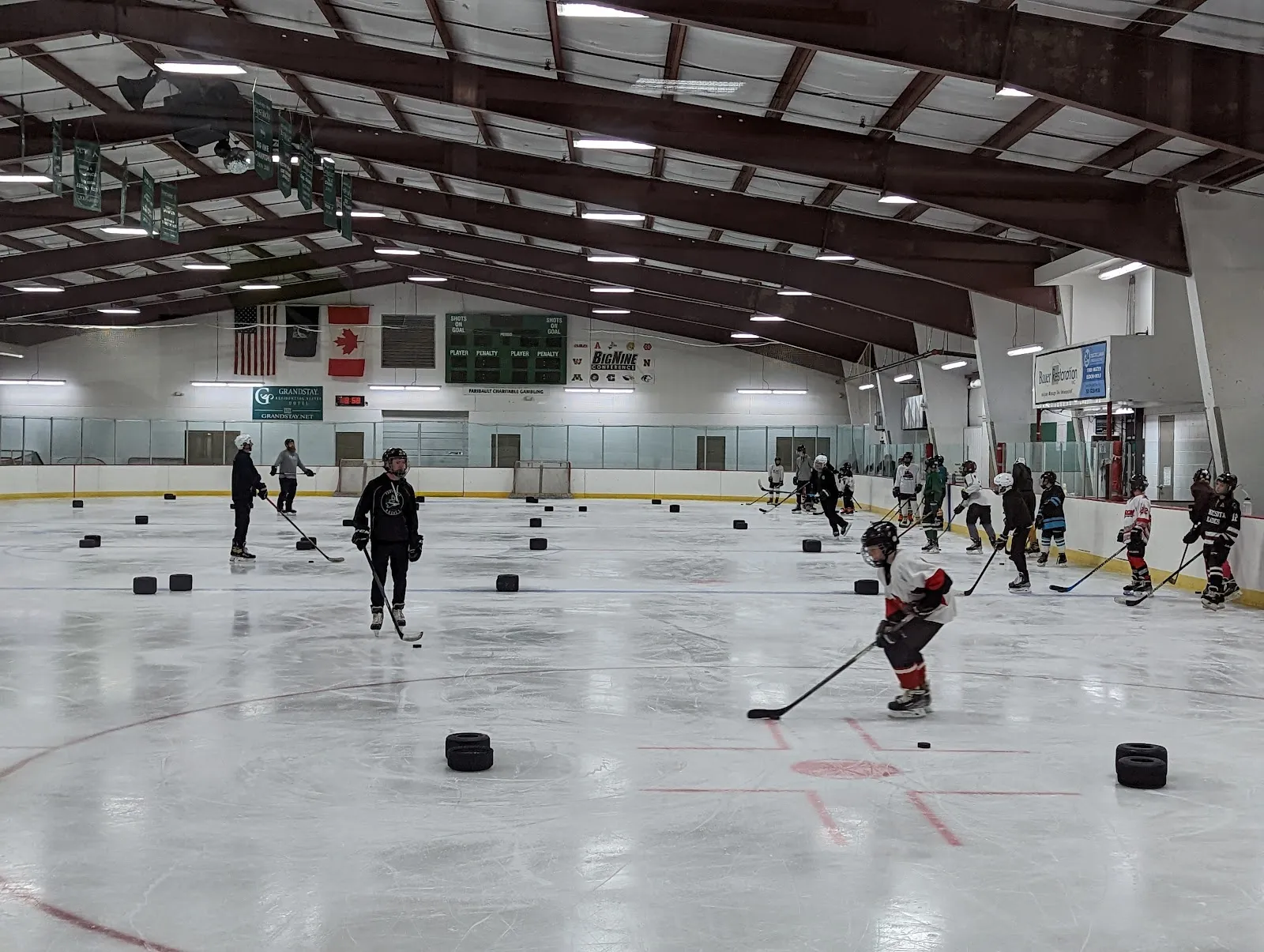 A practice on the sheet seen from the glass, players spread across white ice with pucks scattered and a green-trimmed steel-trussed roof overhead. — Faribault Ice Arena