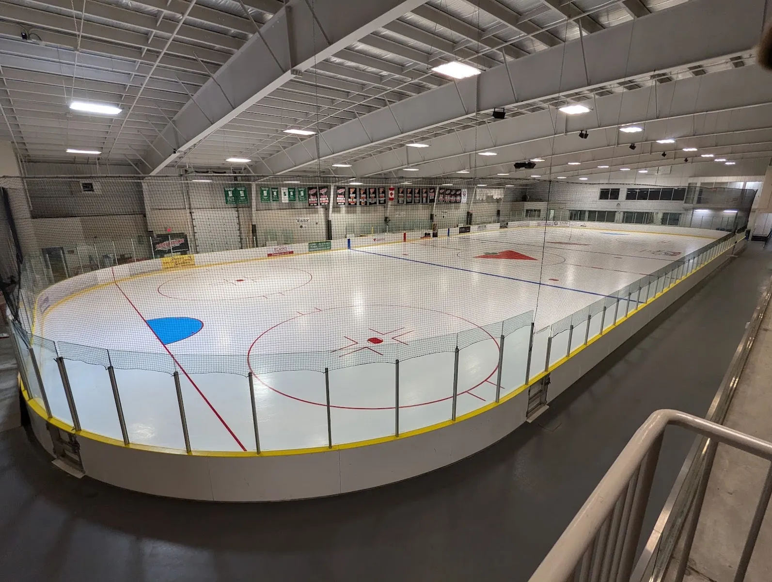 La pista vacía desde las escaleras: hielo recién pulido, zócalos amarillos en las vallas y asientos a lo largo del lado opuesto bajo un techo blanco. — Fenelon Falls Community Centre