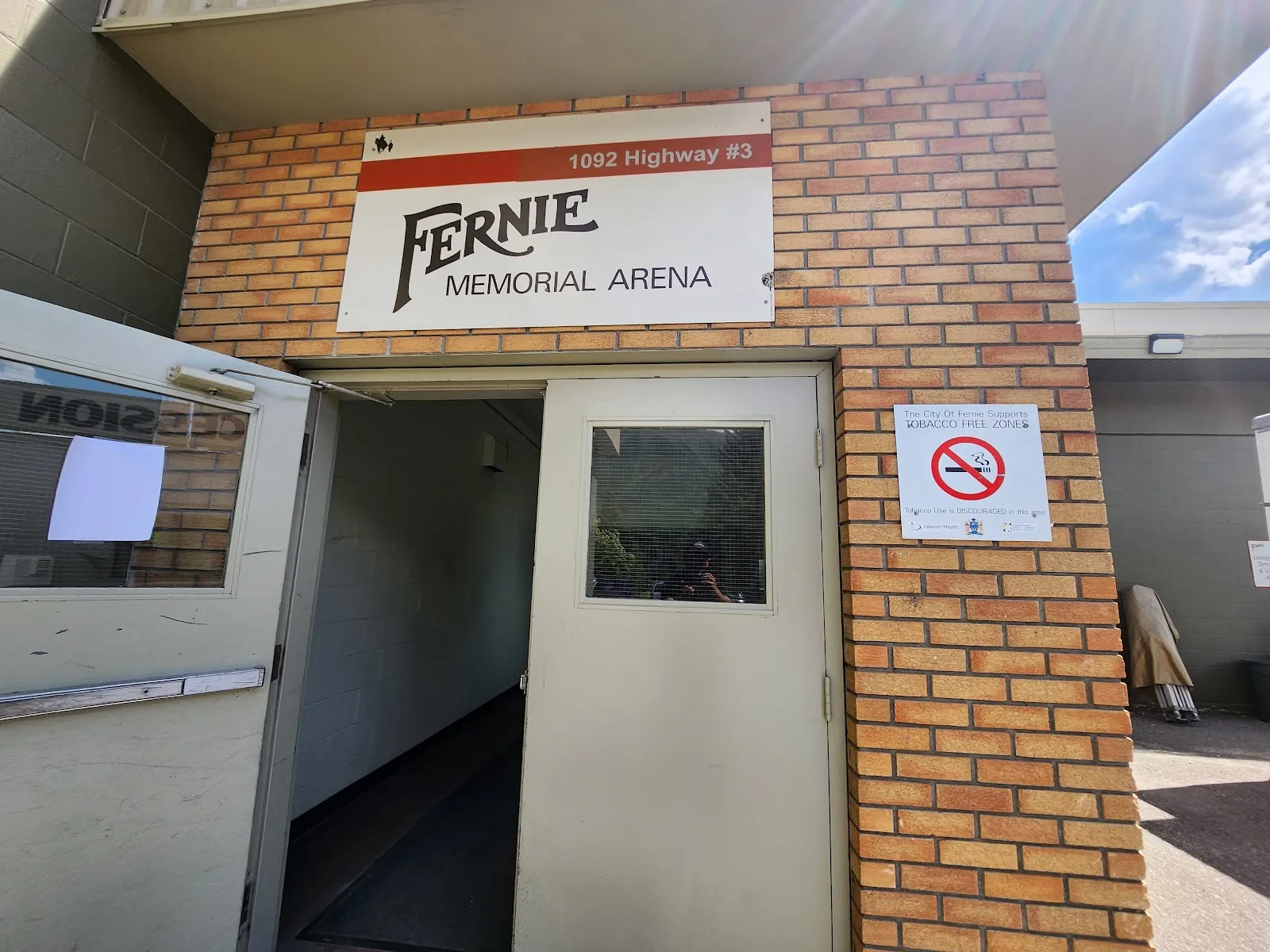 A side entrance of Fernie Memorial Arena in yellow brick with its name and highway address on a white sign, an open door beside it and a no-smoking notice on the wall. — Fernie Memorial Arena
