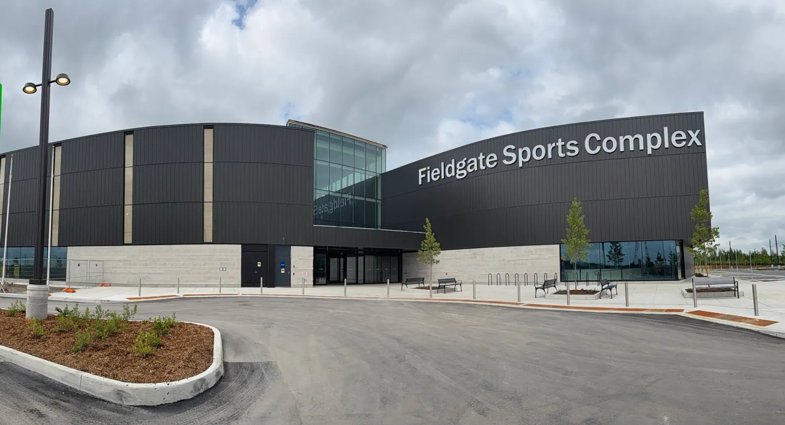 La entrada principal con revestimiento oscuro de Fieldgate Sports Complex, con el nombre en letras grandes en la parte alta del muro, un vestíbulo de vidrio y un camino curvo al frente. — Fieldgate Sports Complex