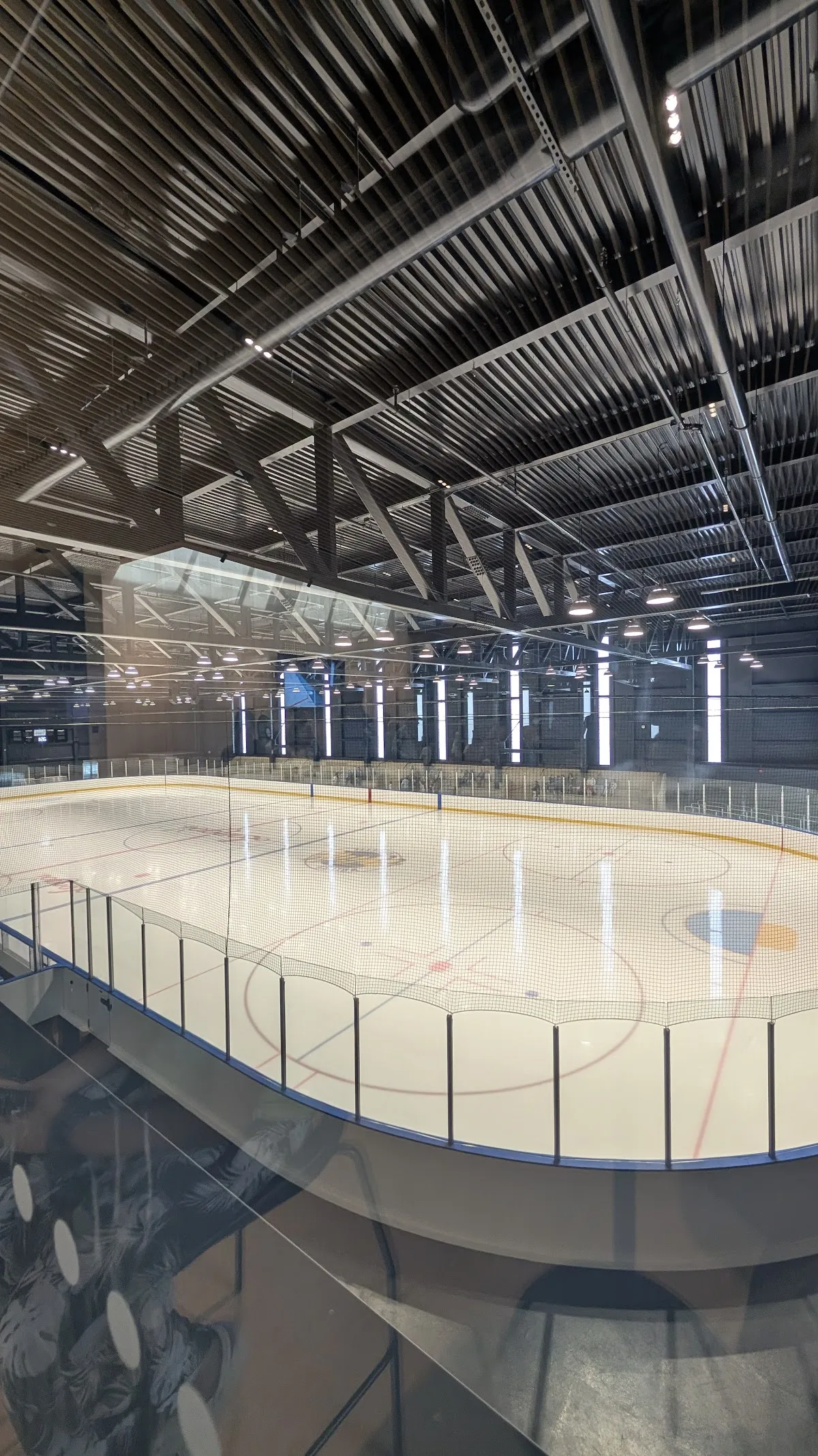Una de las nuevas arenas desde las gradas: toda una pista de hielo bajo un techo de acero expuesto, con cristal a lo largo de las vallas y bancas en el lado cercano. — Fieldgate Sports Complex