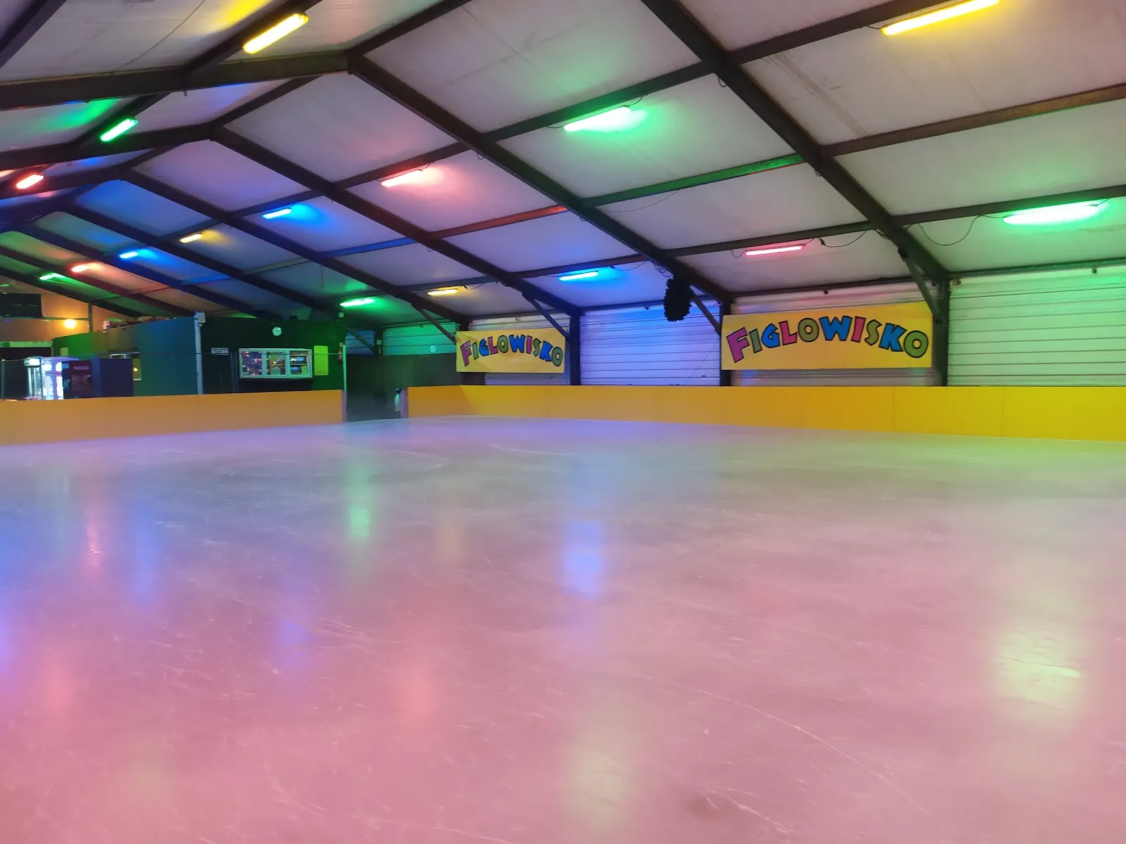 The empty ice of the Figlowisko covered rink in Warsaw Ursynów under its steel roof, lit in colours, with the Figlowisko banners along the boards — Figlowisko Lodowisko 24h