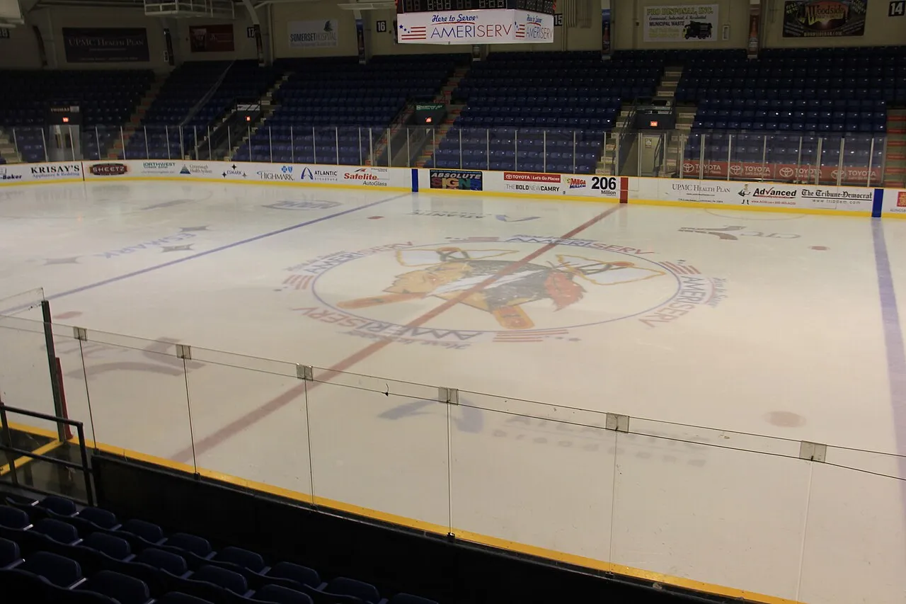 Toda la pista desde un asiento del nivel superior, con el logotipo de hockey de AmeriServ/Tomahawks en el centro de la pista, el cristal y el barandal, y asientos azules vacíos alrededor de la pista. — 1st Summit Arena