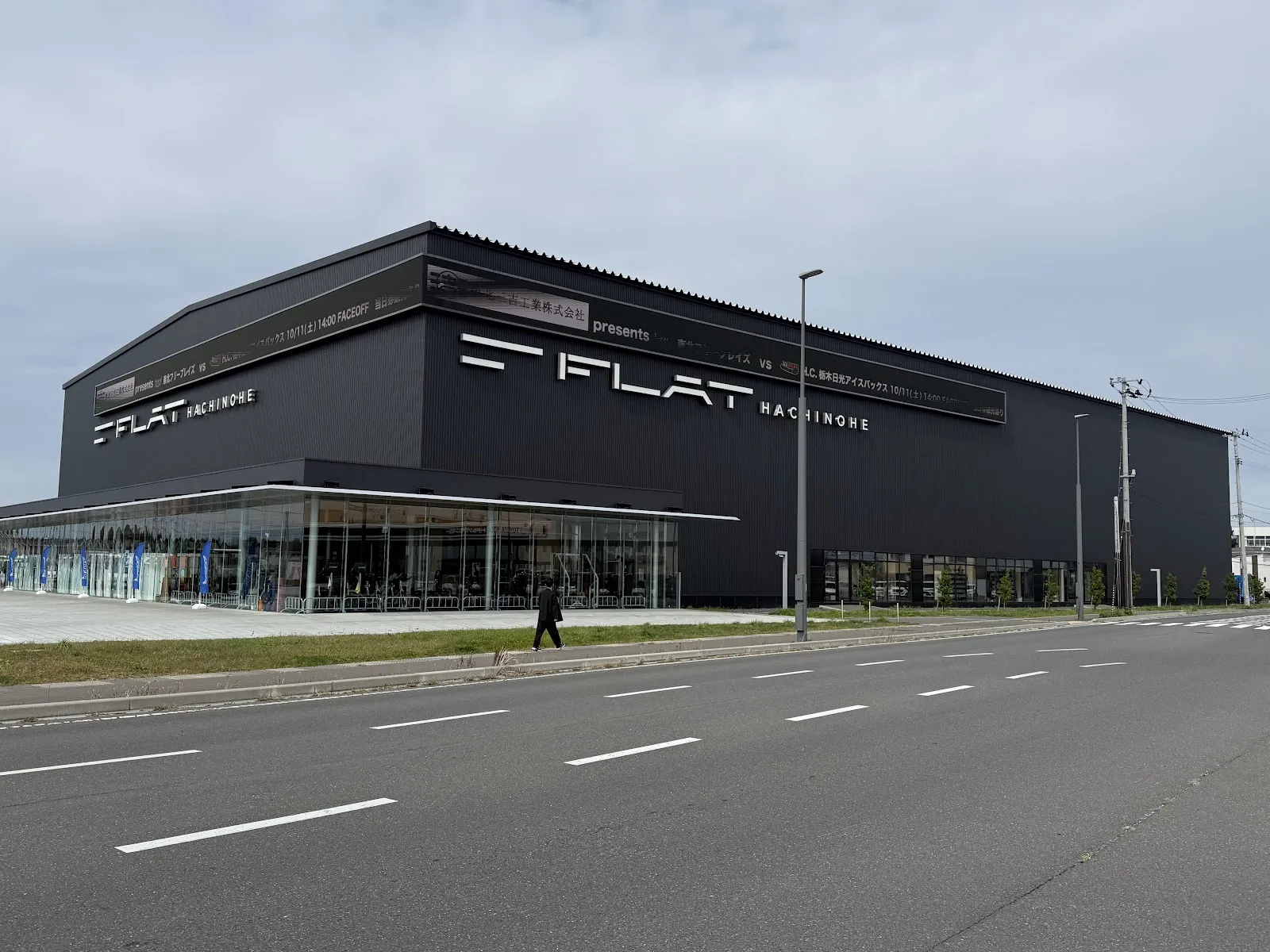 The black exterior of FLAT HACHINOHE seen from the road, with the arena name on the front and a glass entrance hall at street level — FLAT HACHINOHE