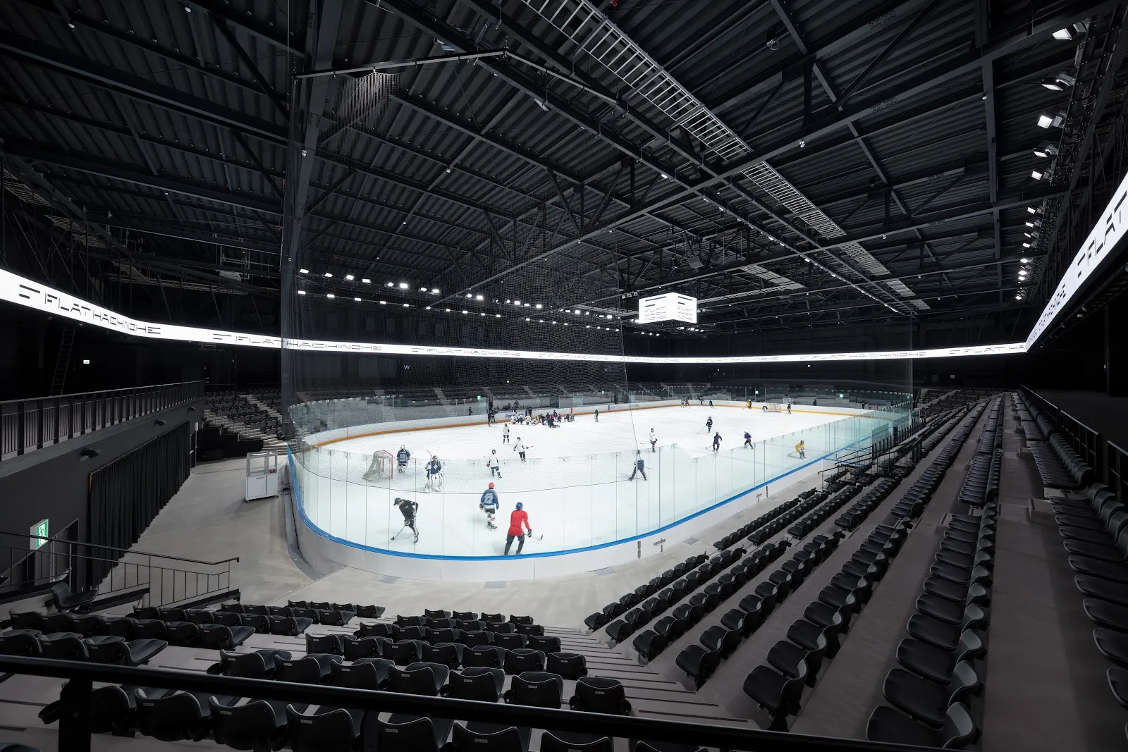 The ice sheet inside FLAT HACHINOHE seen from the stands, with a few small skaters on the ice and the ring of LED screens above — FLAT HACHINOHE