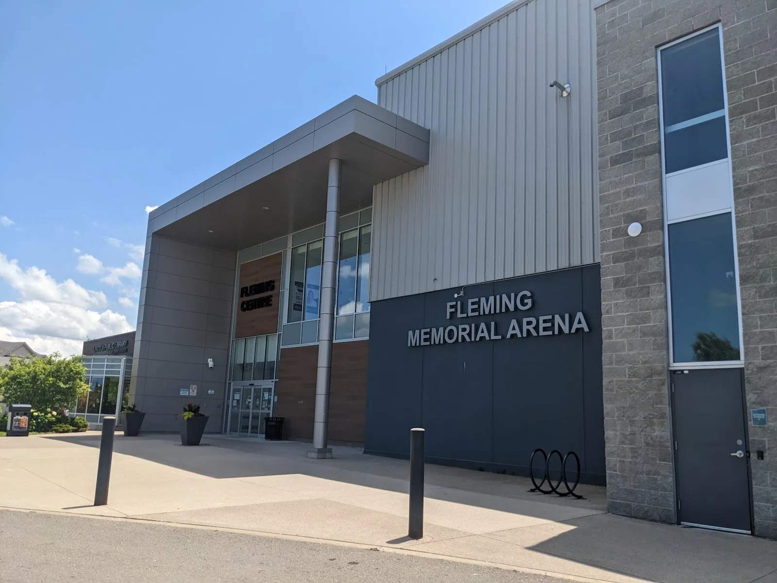 The Fleming Memorial Arena entrance on a sunny day: grey metal cladding and stone, with the arena name in large letters beside the glass lobby. — Fleming Memorial Arena