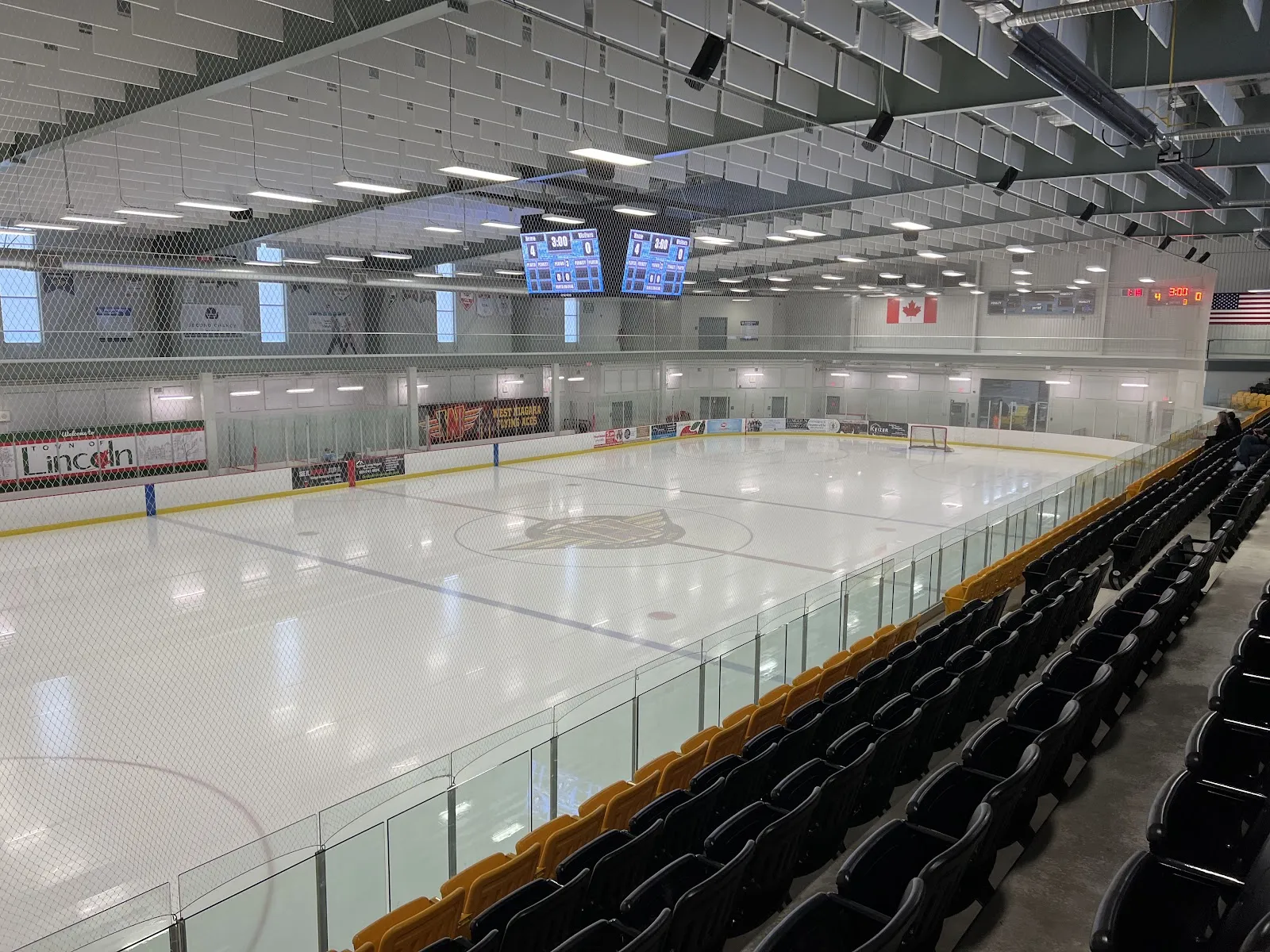 The Fleming Memorial Arena ice from the stands, with rows of black and gold seats, scoreboards over centre ice and Town of Lincoln boards. — Fleming Memorial Arena