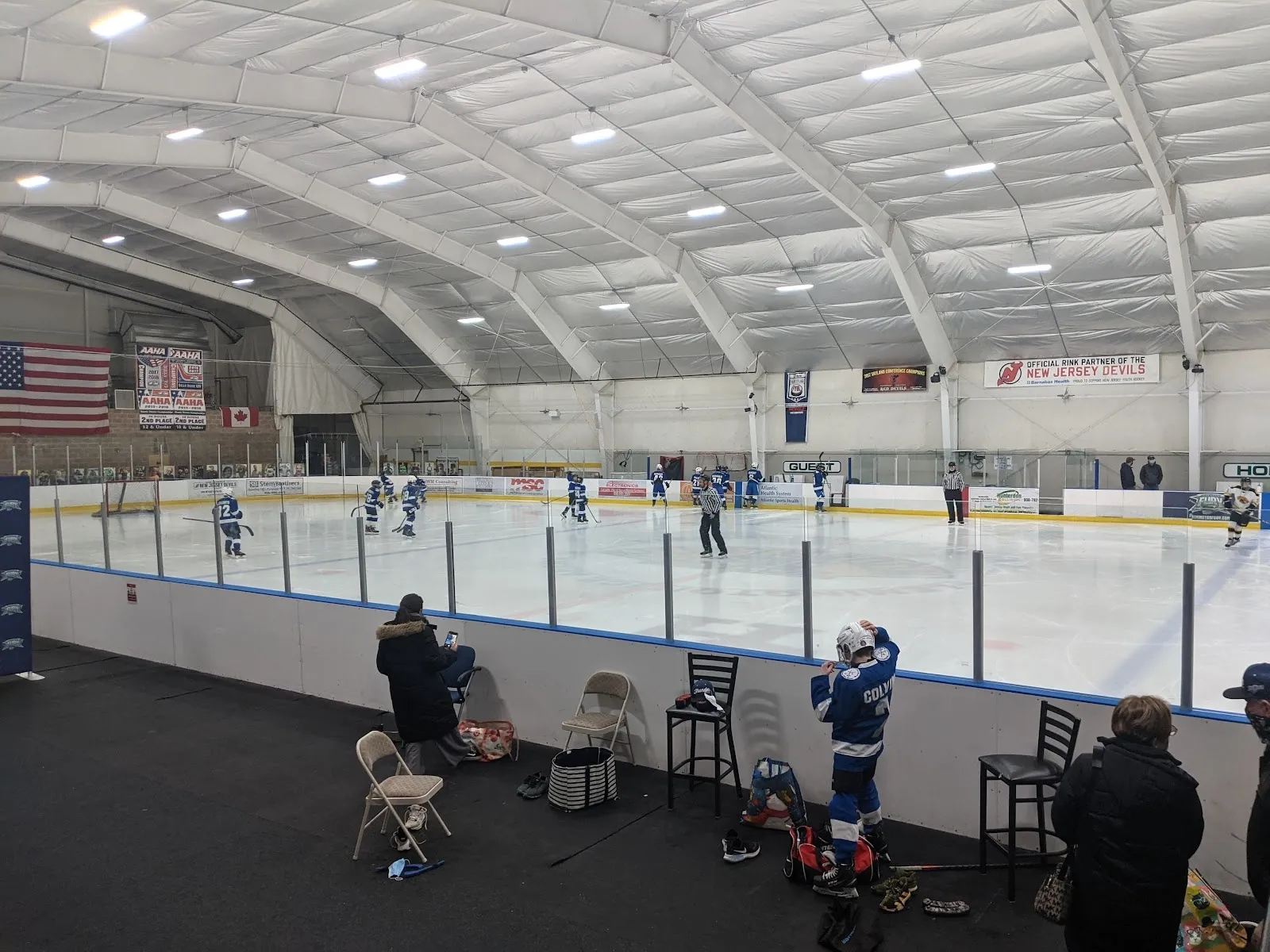 Una vista amplia a lo largo del vidrio con unos cuantos patinadores y un entrenador junto a las vallas cercanas, un techo blanco curvo y una bandera de Estados Unidos sobre la pared del fondo. — Flemington Ice Arena