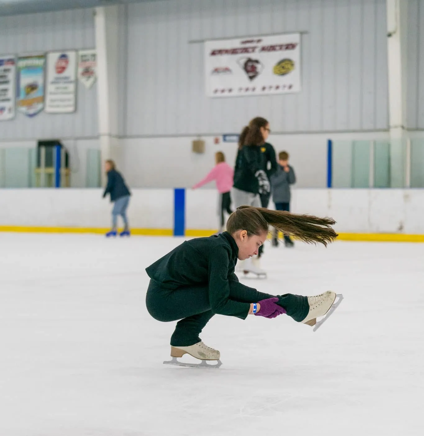 Una patinadora artística en un giro sobre el hielo, con patinadores del público a lo largo de las vallas detrás de ella y banderines de hockey en la pared. — Flight Adventure Park Irmo (ice rink)